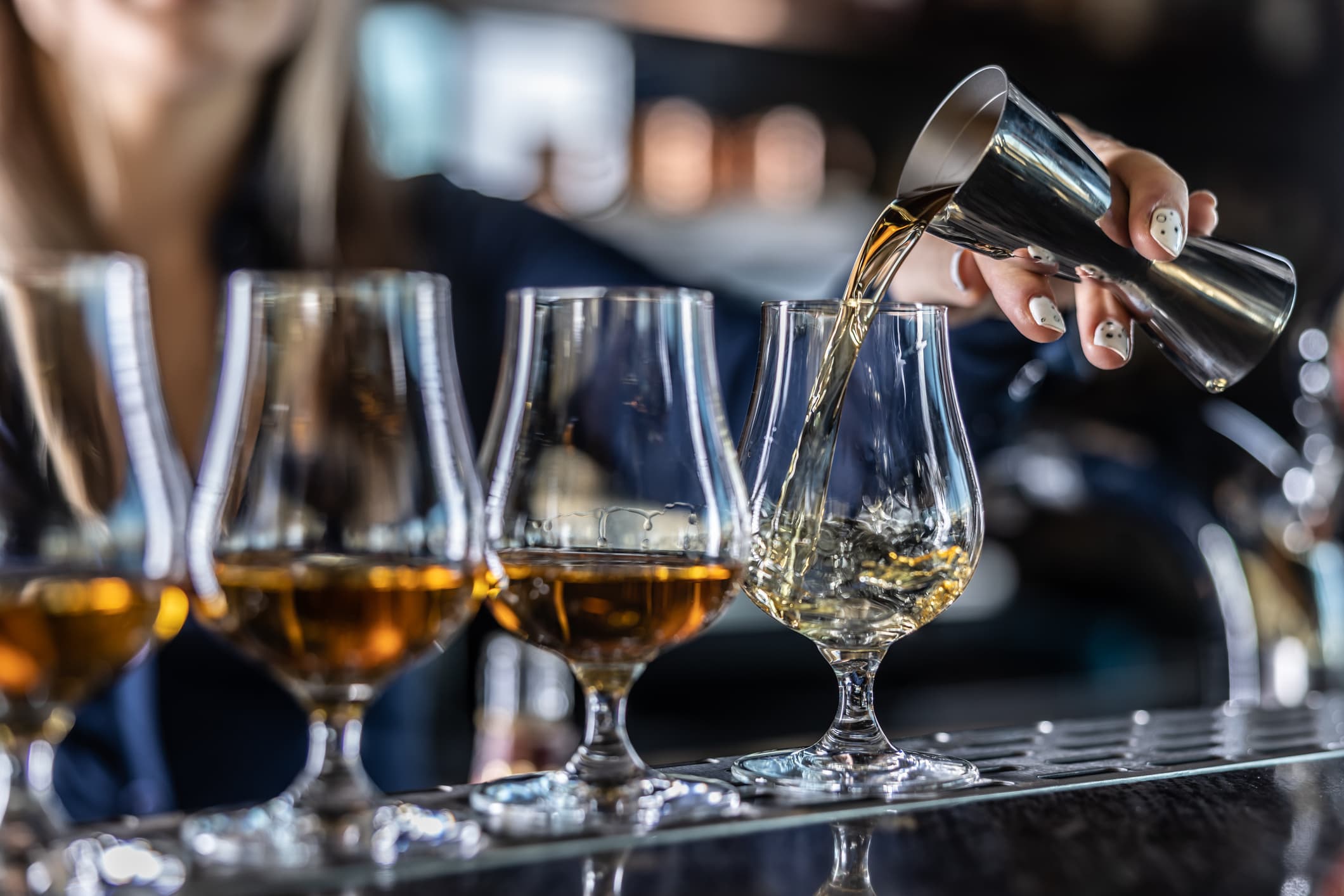 Young female bartender professionally pours rum or brandy from steel jigger into glass at bar.